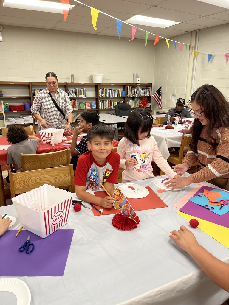 Children create colorful crafts at a classroom table while adults assist; festive bunting, library shelves, and bright artwork create a joyful mood.