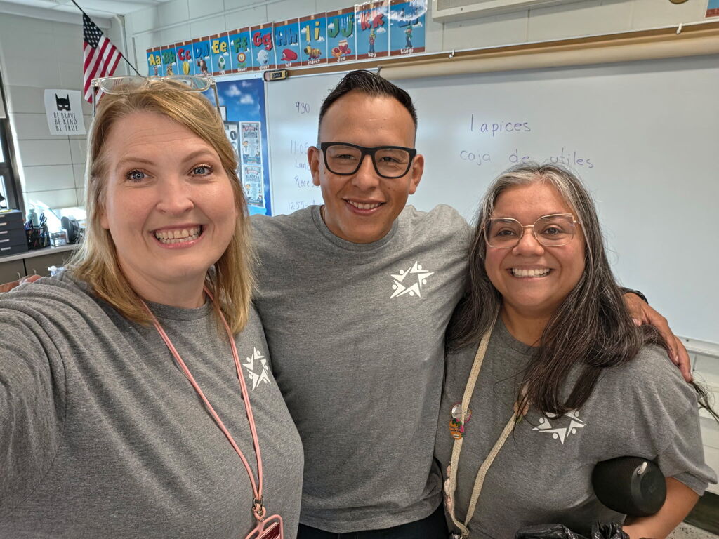Three smiling teachers pose together in a classroom, wearing matching gray shirts; a U.S. flag and colorful student artwork appear behind them.