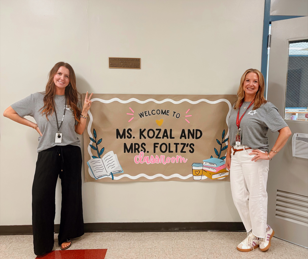 Two smiling teachers pose beside a colorful “Welcome to Ms. Kozal and Mrs. Foltz’s classroom” banner in a school hallway.