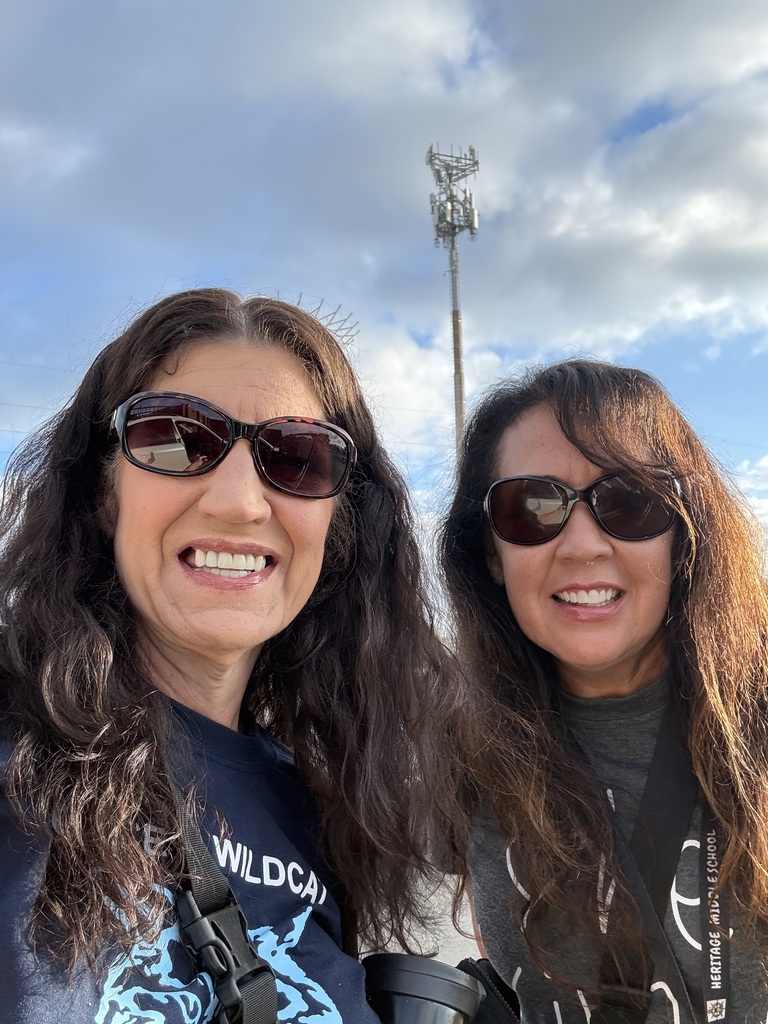 Two smiling women in sunglasses pose outdoors beneath a blue, cloudy sky, with a cell tower behind them; cheerful, friendly mood.