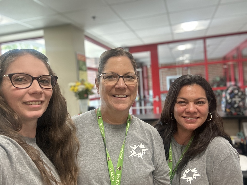 Three smiling women wearing glasses and gray shirts pose together indoors, with green lanyards and a bright red retail or community space behind them.