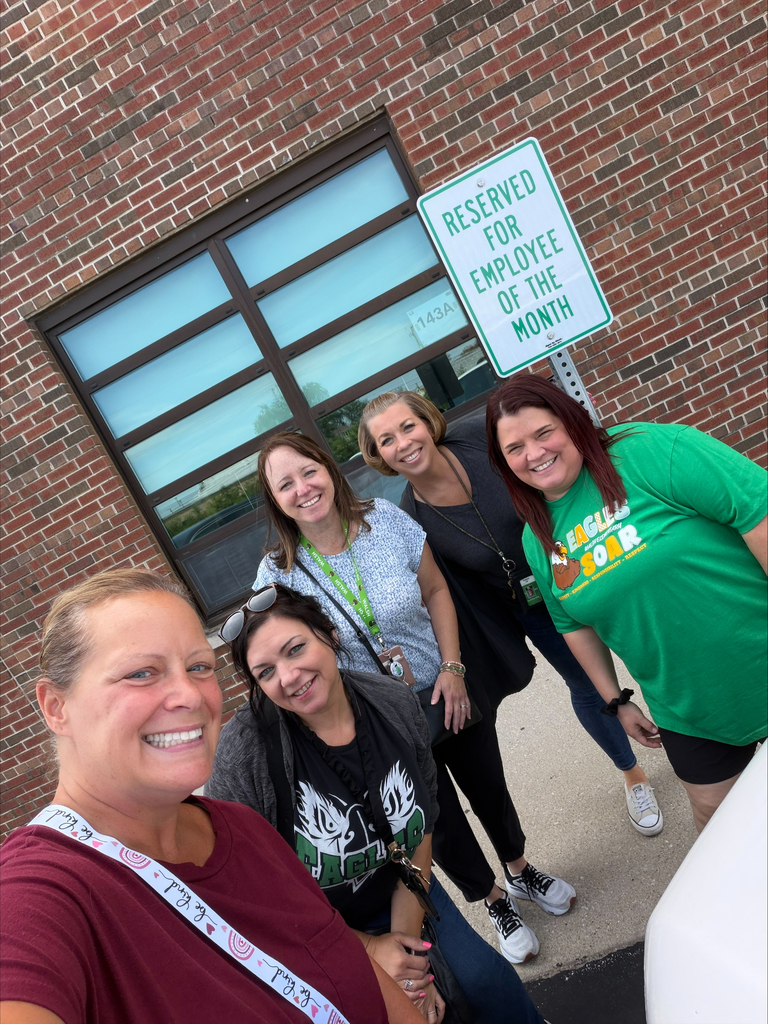 Cheerful group selfie of five women in casual clothes outside a brick building, smiling beneath a “Reserved for Employee of the Month” sign.