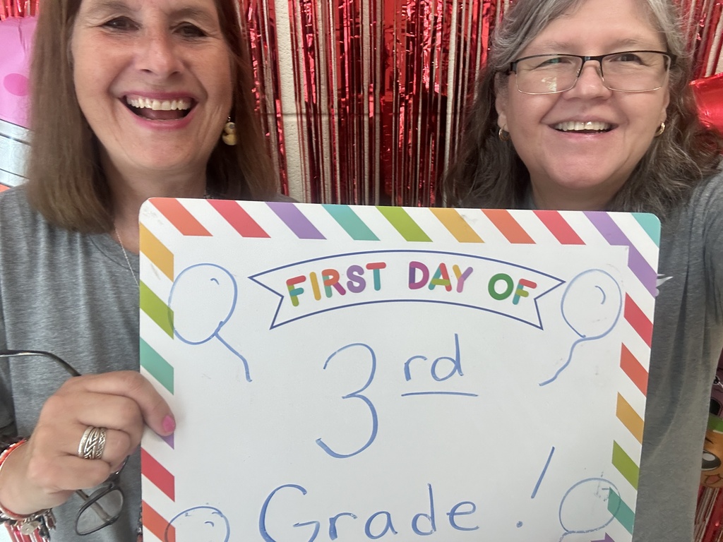 Two smiling women hold a colorful “First Day of 3rd Grade” sign before shimmering red tinsel, creating a cheerful school celebration.