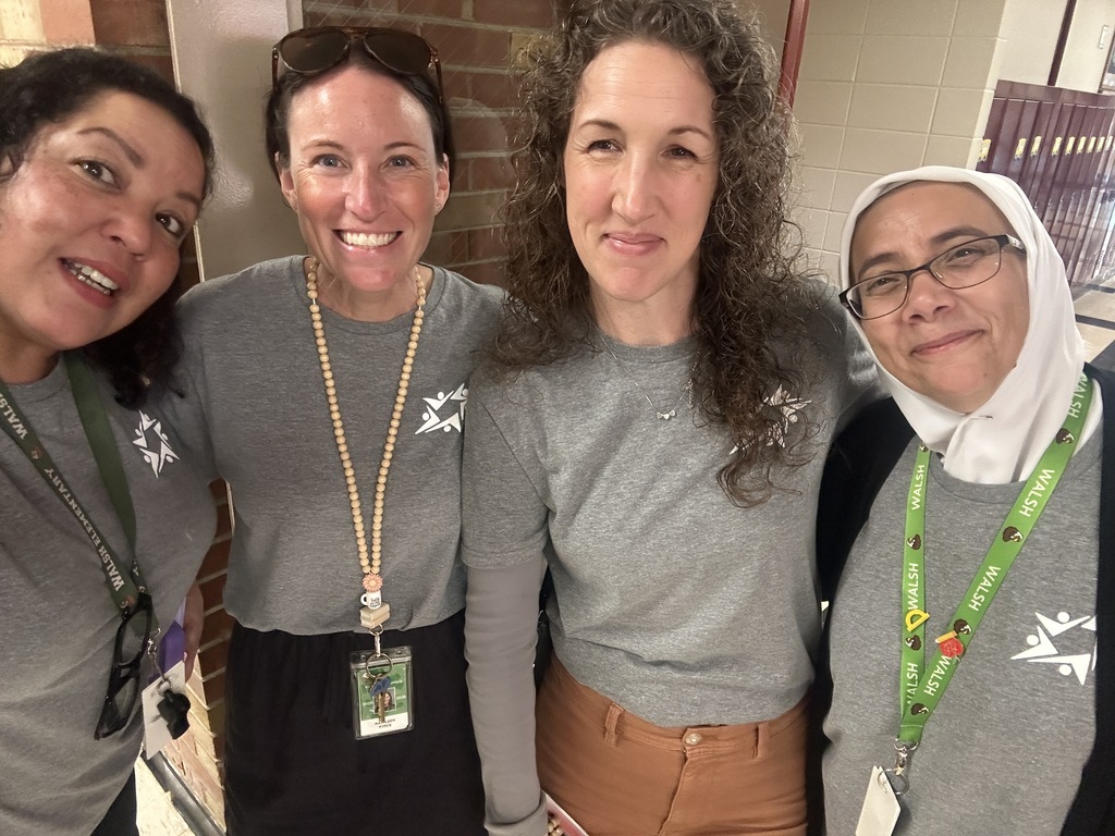 Four smiling women pose for a friendly selfie indoors, wearing gray shirts and lanyards against a brick wall, conveying warmth and camaraderie.
