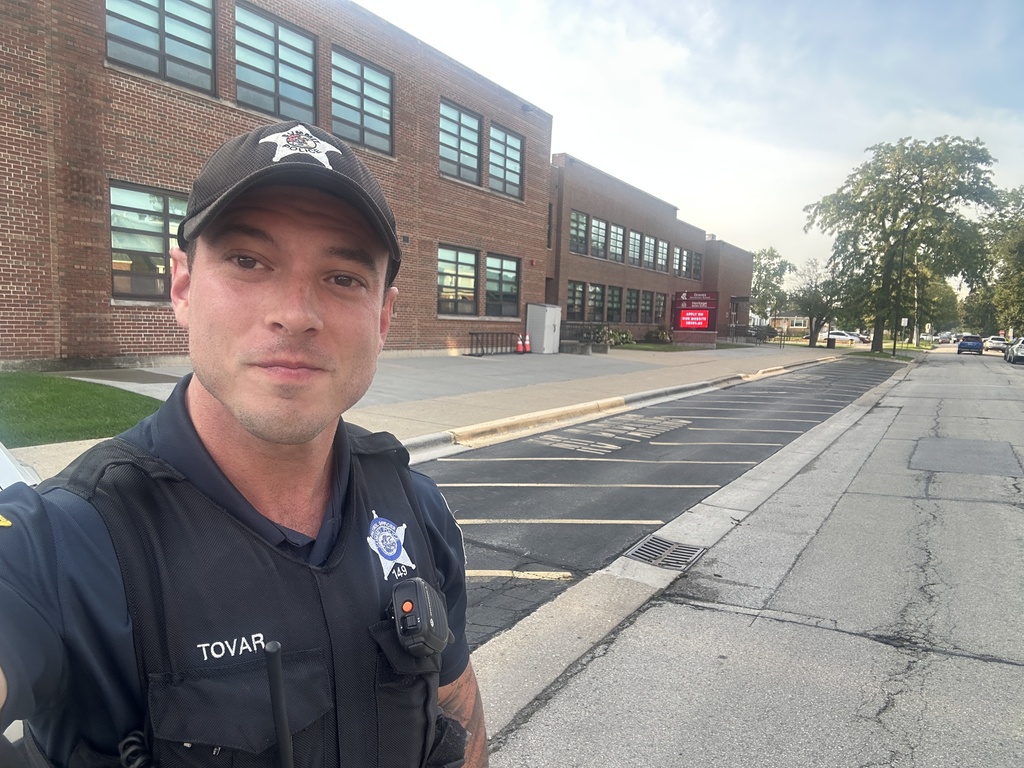 Uniformed officer Tovar takes a selfie outside a brick school building, with empty parking spaces, trees, and a sunny street behind him.