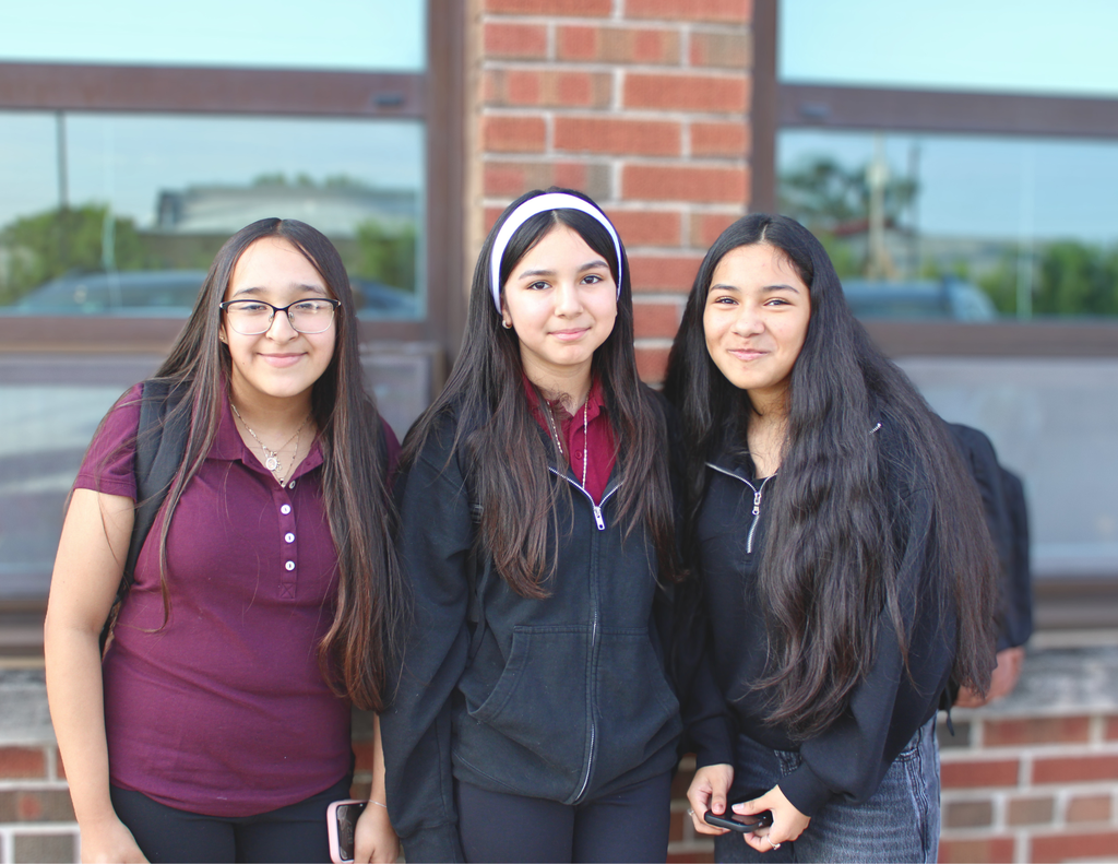 Three smiling teenage girls pose together outside a brick building, wearing casual clothes and backpacks, conveying friendship and warmth.