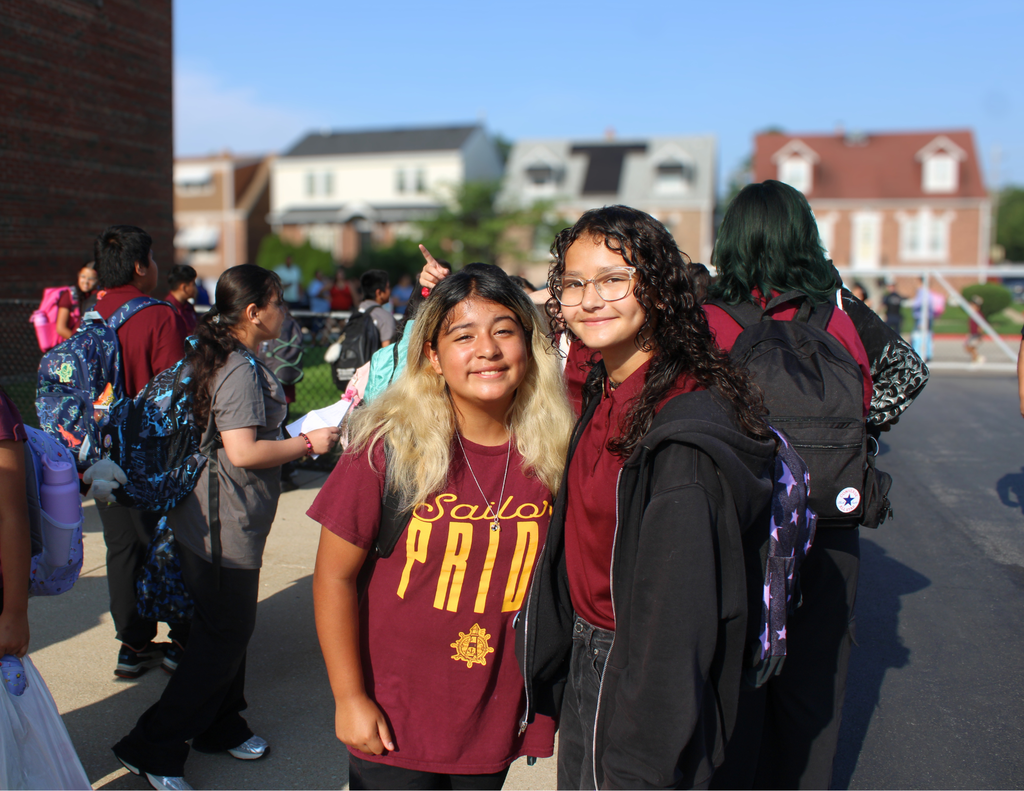 Two smiling students pose together at a school event, one wearing a maroon “Sailor Pride” shirt, amid classmates and suburban homes.
