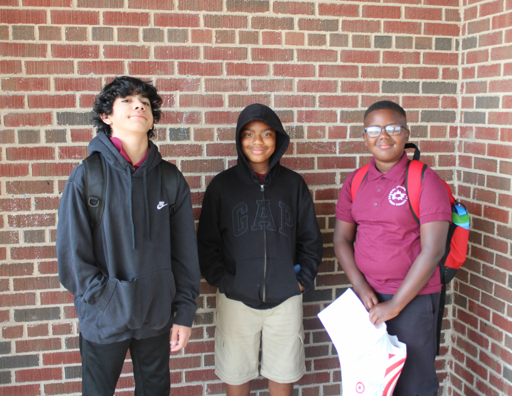 Three diverse students smile in front of a brick wall, wearing backpacks; one holds papers, creating a friendly, relaxed school portrait.