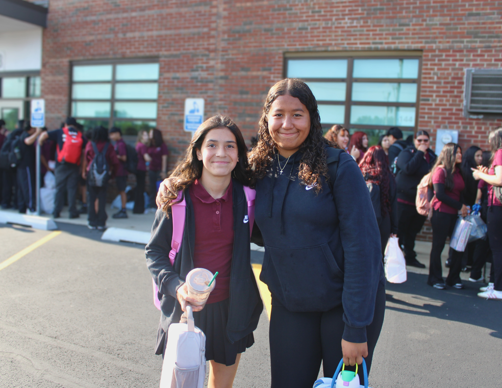 Two smiling students pose outside a brick school building, one holding a drink and suitcase, while a crowd gathers behind them.