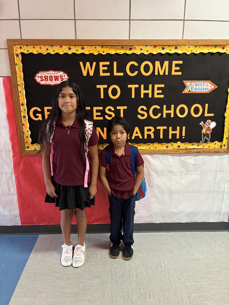 Two smiling schoolchildren pose before a colorful bulletin board reading “Welcome to the Greatest School on Earth!” in a cheerful classroom.