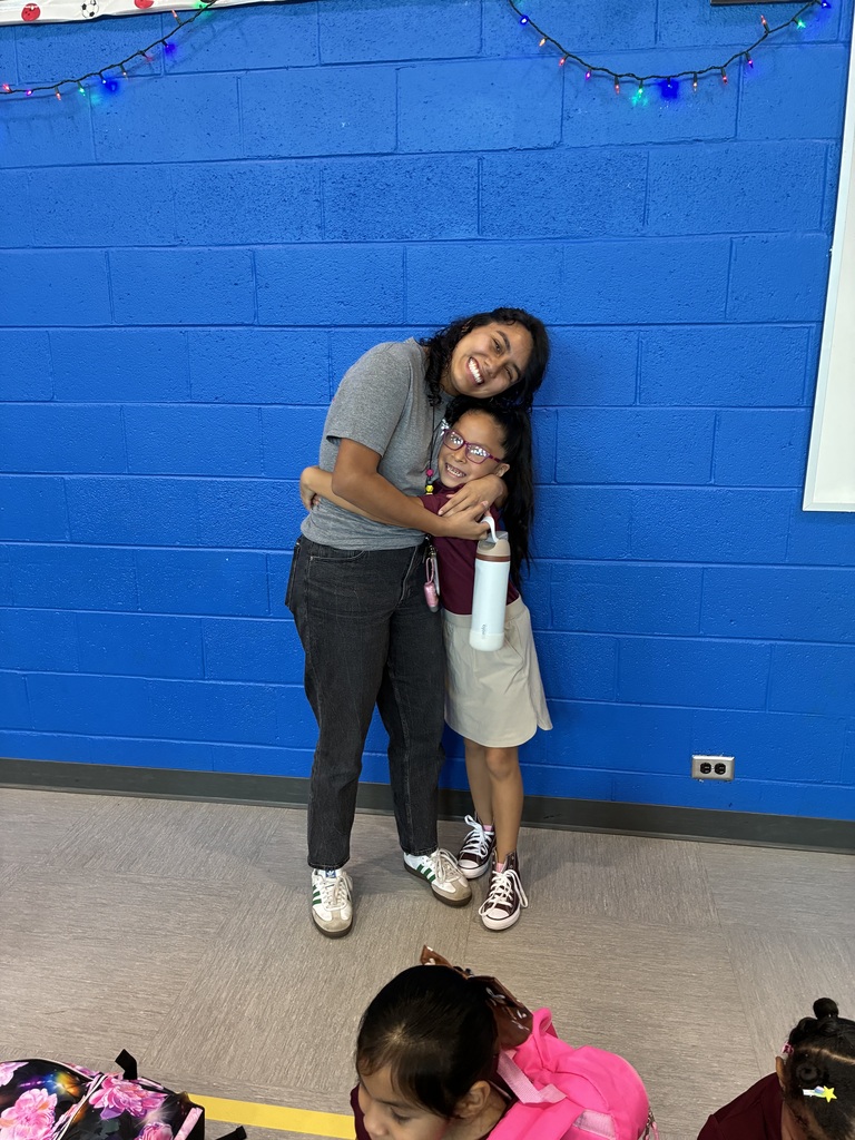 Two smiling girls hug in front of a bright blue cinderblock wall with string lights, while a child in pink sits in the foreground.