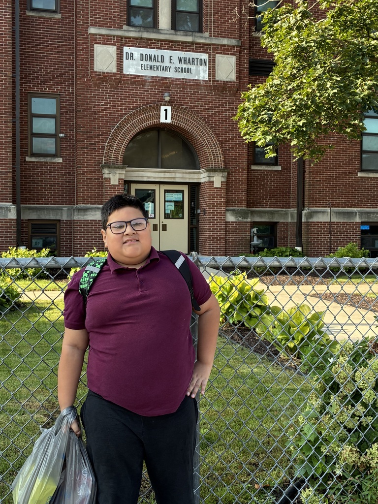 Smiling student poses outside Dr. Donald E. Wharton Elementary School, wearing a maroon shirt and backpack behind a chain-link fence.