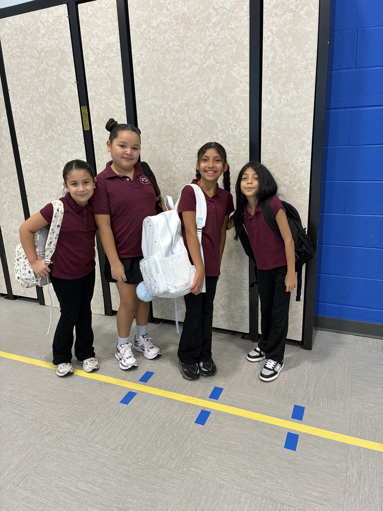 Four smiling schoolgirls in maroon school uniforms pose with backpacks in a hallway beside blue and yellow floor markings, creating a cheerful scene.