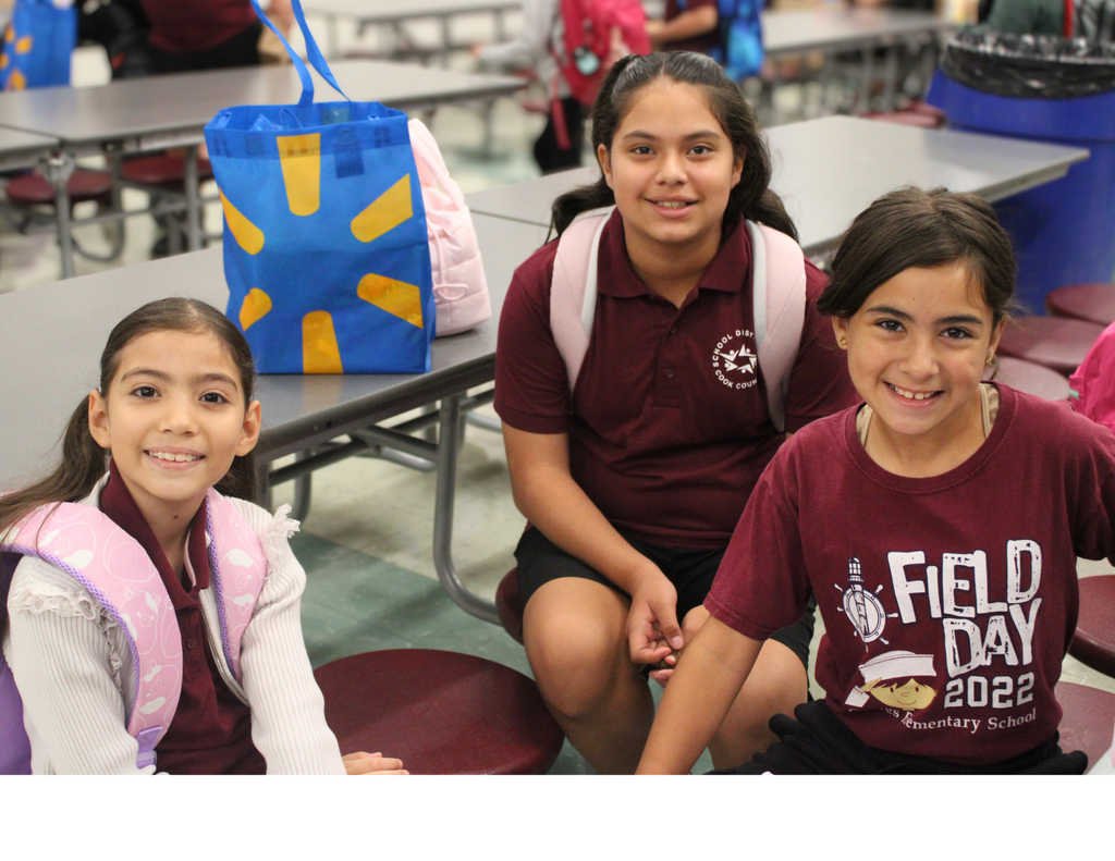 Three smiling schoolgirls pose together in a school cafeteria, wearing backpacks and maroon shirts, creating a cheerful, friendly scene.