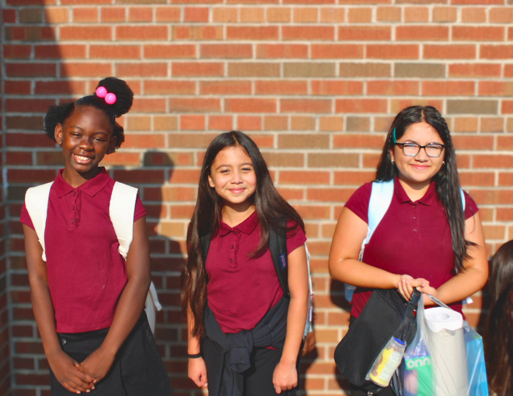 Three smiling schoolgirls in burgundy uniforms stand outside a red brick school wall, carrying backpacks and a reusable shopping bag.