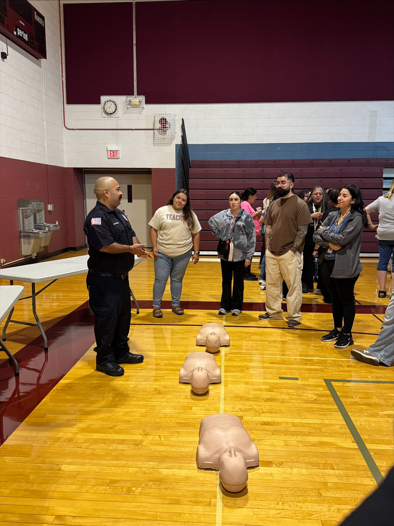 A uniformed instructor demonstrates CPR in a school gym as adults watch mannequins arranged on the polished basketball court.