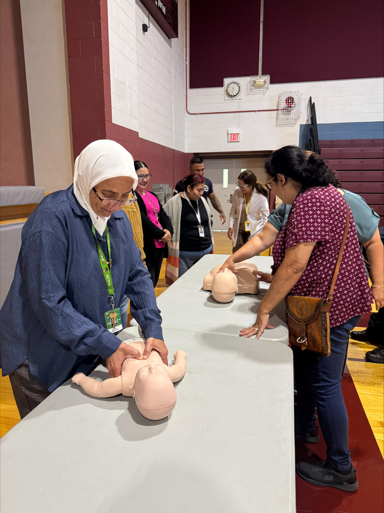 Adults practice CPR on infant mannequins at a community safety class in a gym, with an instructor guiding hands-on group training.