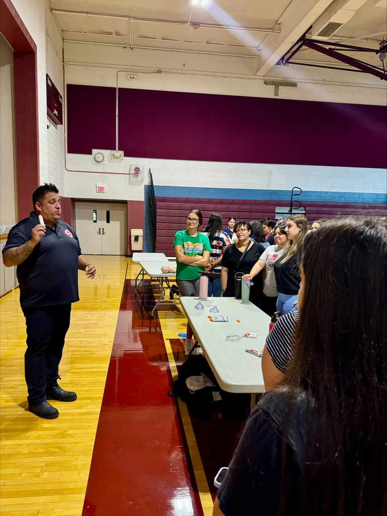Speaker addresses a seated group beside a long table in a school gym, with colorful bleachers and polished wood floors creating an attentive, upbeat mood.