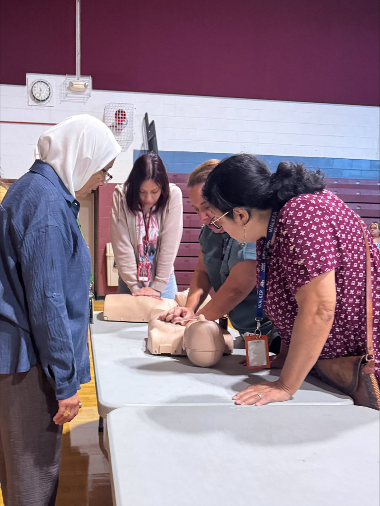Women in a hands-on CPR training session press on a mannequin’s chest at a table, watched by colleagues in a classroom; focused, collaborative mood.
