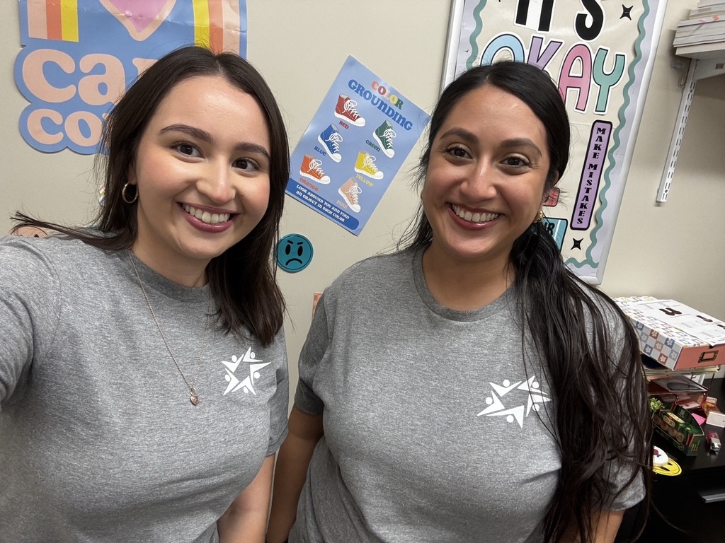 Two smiling women in matching gray shirts pose for a selfie in a colorful classroom or office, surrounded by cheerful posters and supplies.