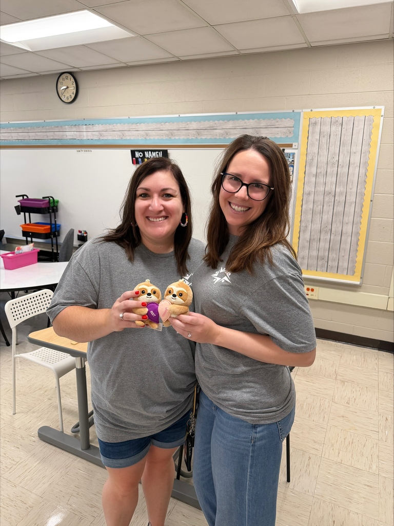 Two smiling women in matching gray shirts hold colorful plush toys in a classroom, posing cheerfully beneath a clock and whiteboard.
