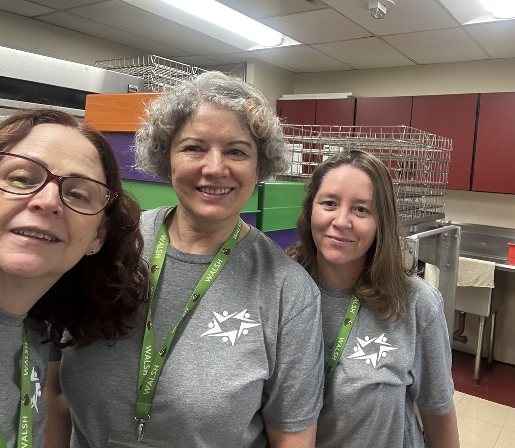 Three smiling women in matching gray shirts and green lanyards pose for a selfie in a bright workplace, with colorful shelves behind them.