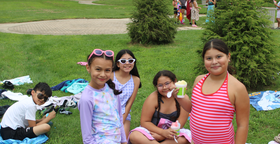Four girls pose on a grassy park lawn, smiling and holding drinks; two wear sunglasses, while people, towels, and trees fill the background.
