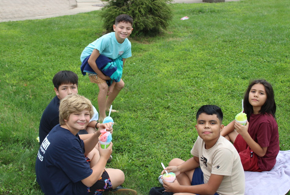 Five children sit and kneel on a green lawn, smiling while enjoying colorful ice cream treats on a bright, playful afternoon.