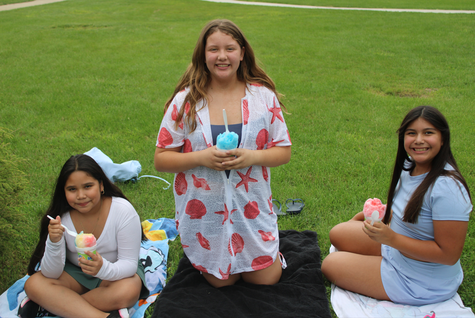 Three smiling girls sit on blankets in a grassy park, holding colorful shaved ice treats during a cheerful outdoor picnic.