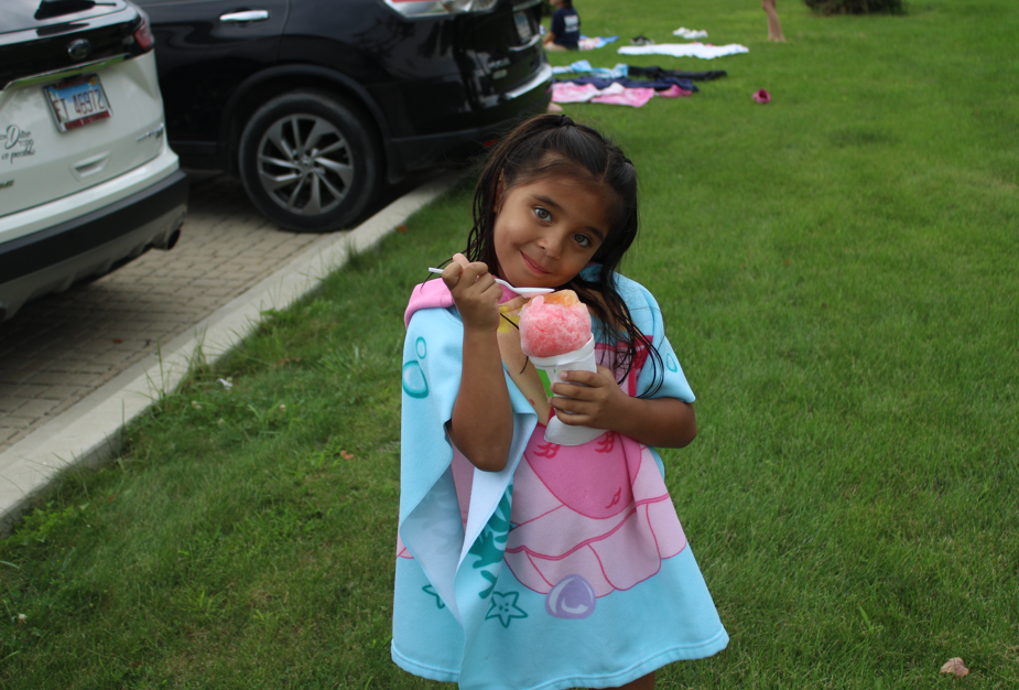 Smiling young girl in a colorful mermaid towel holds a pink ice cream on a grassy lawn, with parked cars and scattered towels behind her.