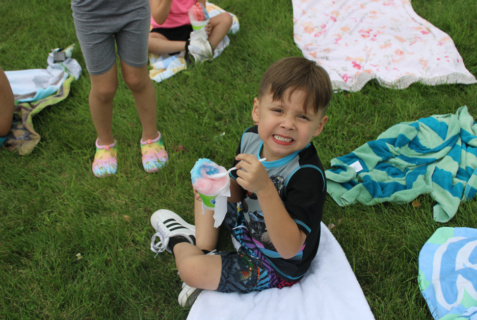 Smiling young boy sits on a blanket in the grass, holding a colorful frozen treat, while other children and picnic blankets surround him.