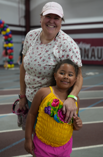 Smiling woman in a pink cap and floral shirt hugs a young girl in a bright yellow embroidered dress at an indoor gym, conveying warmth and joy.