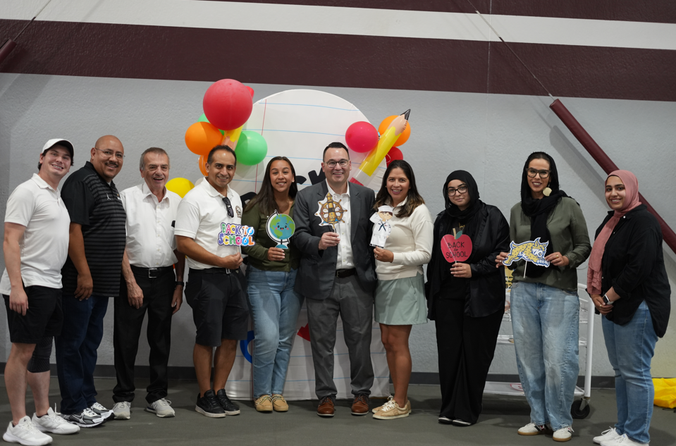 A diverse group poses cheerfully at an indoor event, holding trophies and colorful decorations, with balloons and a branded backdrop behind them.