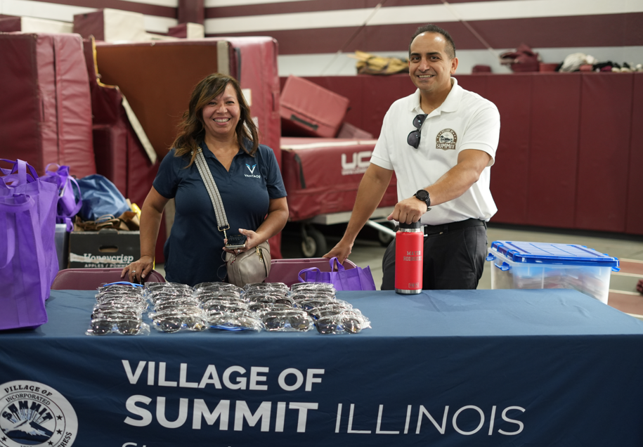 Smiling community volunteers stand behind a Village of Summit, Illinois table with purple bags and wrapped items in a gym, creating a welcoming tone.