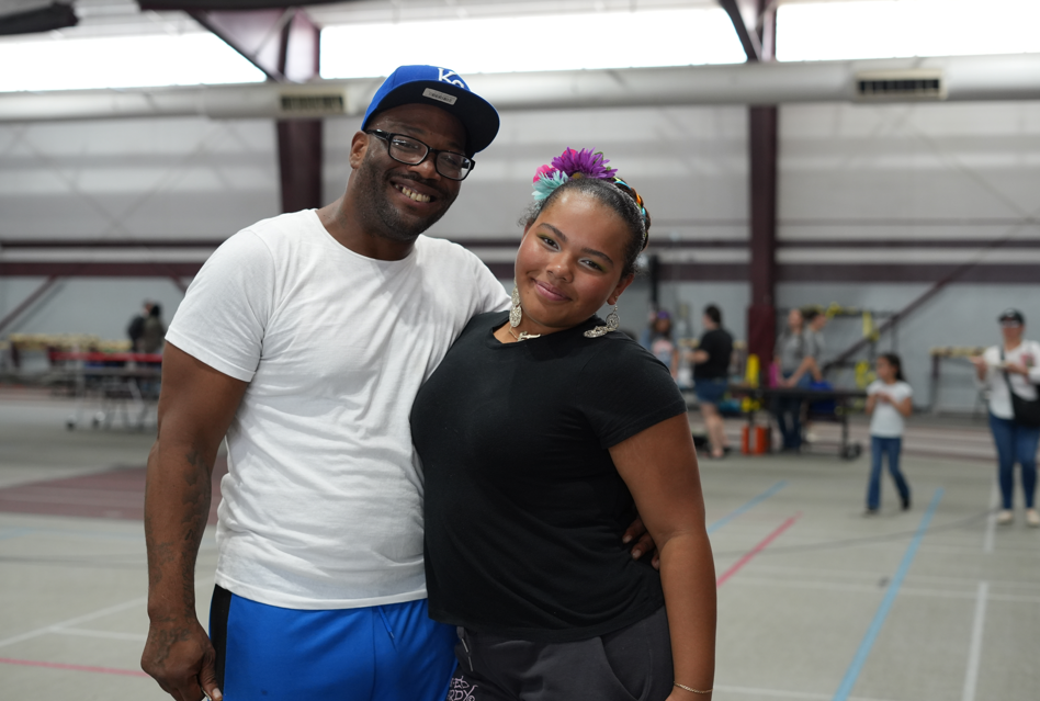 Smiling couple pose together in a brightly lit indoor gym, with colorful court lines and people in the background creating a friendly atmosphere.