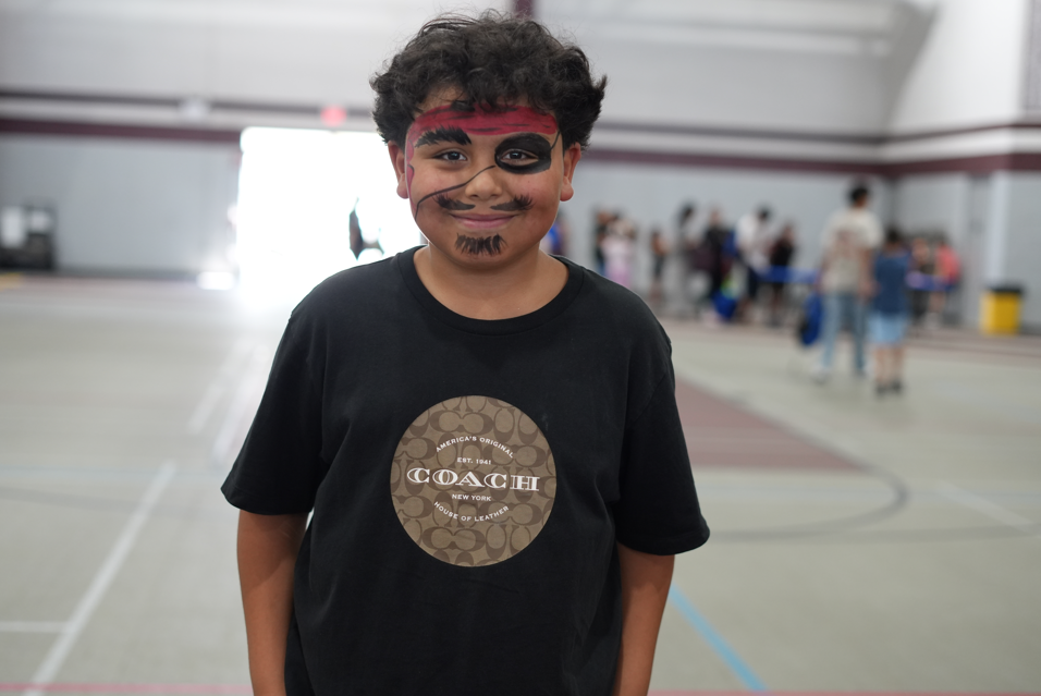 Smiling child with black-and-red face paint wears a black Coach T-shirt in a bright gym, with blurred people and court lines behind him.