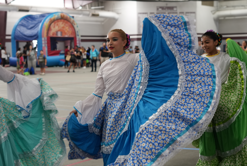 Traditional folkloric dancers in colorful blue and green dresses perform indoors, lifting flowing skirts in a lively cultural celebration.