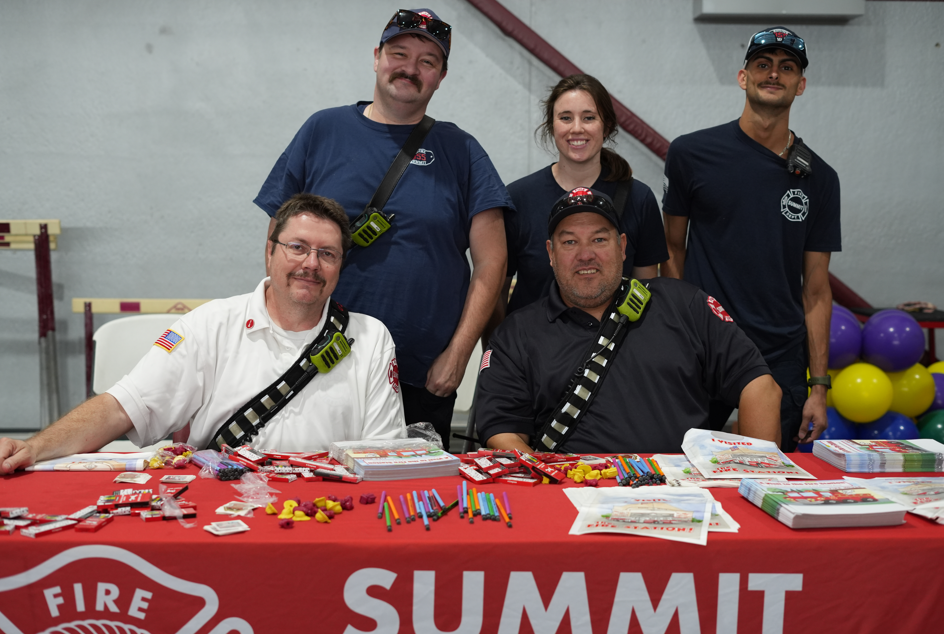 Five smiling firefighters pose behind a red Fire Summit table covered with brochures and giveaways, with colorful balloons in the background.