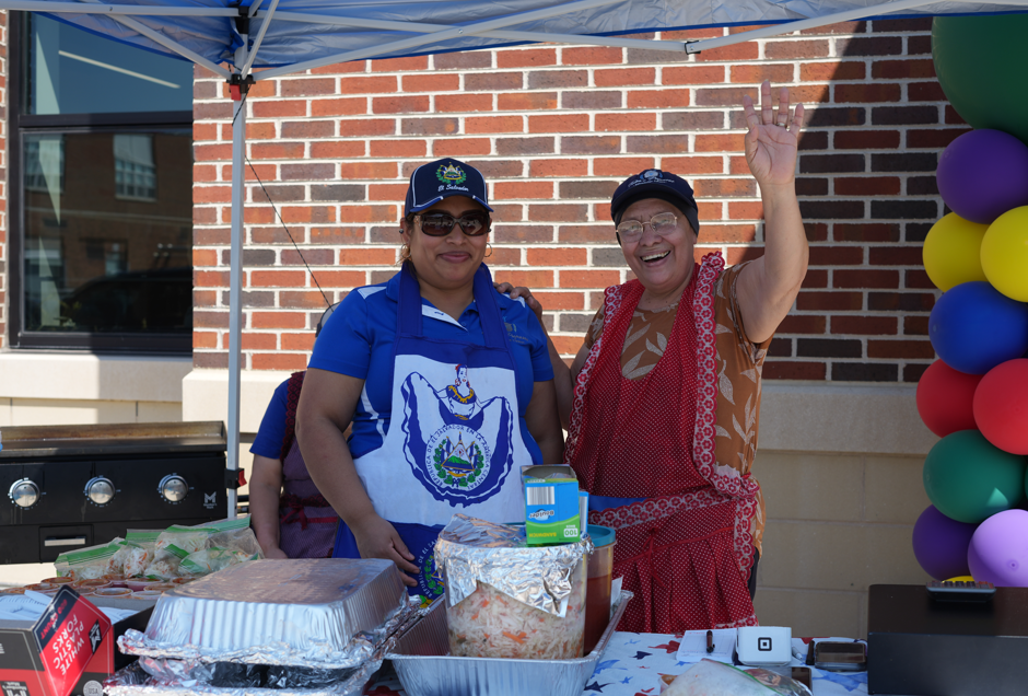 Two smiling women pose at a community food table beneath a canopy, with barbecue trays, colorful balloons, and a brick wall behind them.