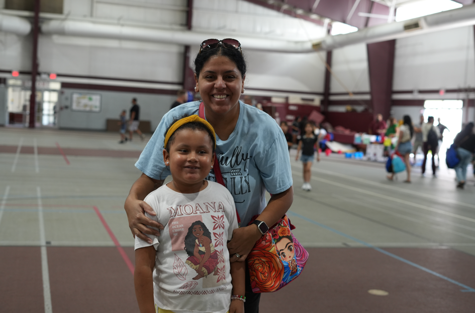 Smiling mother and young child pose together in an indoor gym, with sports courts, people, and colorful event booths behind them.