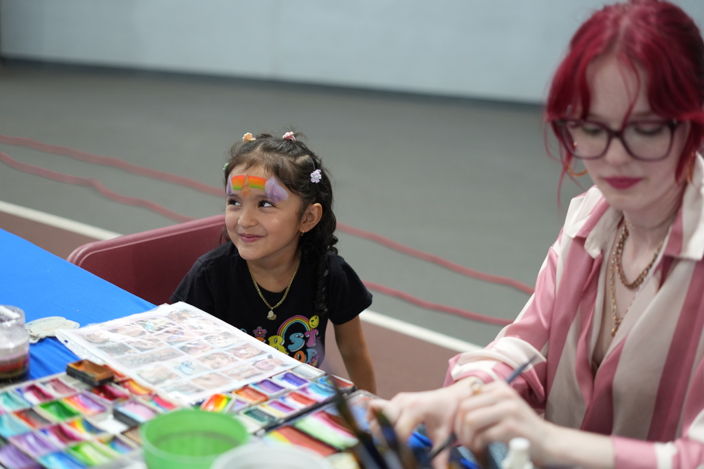 Smiling child and red-haired woman in glasses create art together at a colorful craft table, surrounded by paints and supplies.