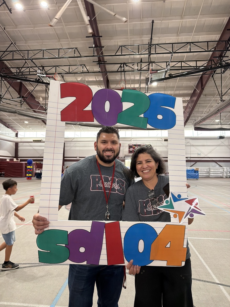 Smiling man and woman pose behind a colorful “2025” photo frame at an indoor event, with a star decoration and spacious gym venue behind them.