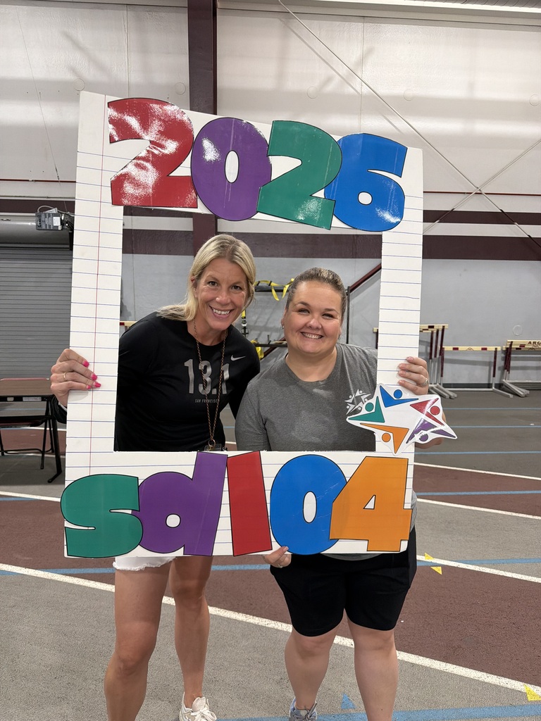 Two smiling women hold a colorful “2024” sign in an indoor gym, conveying joy and celebration at a community event.