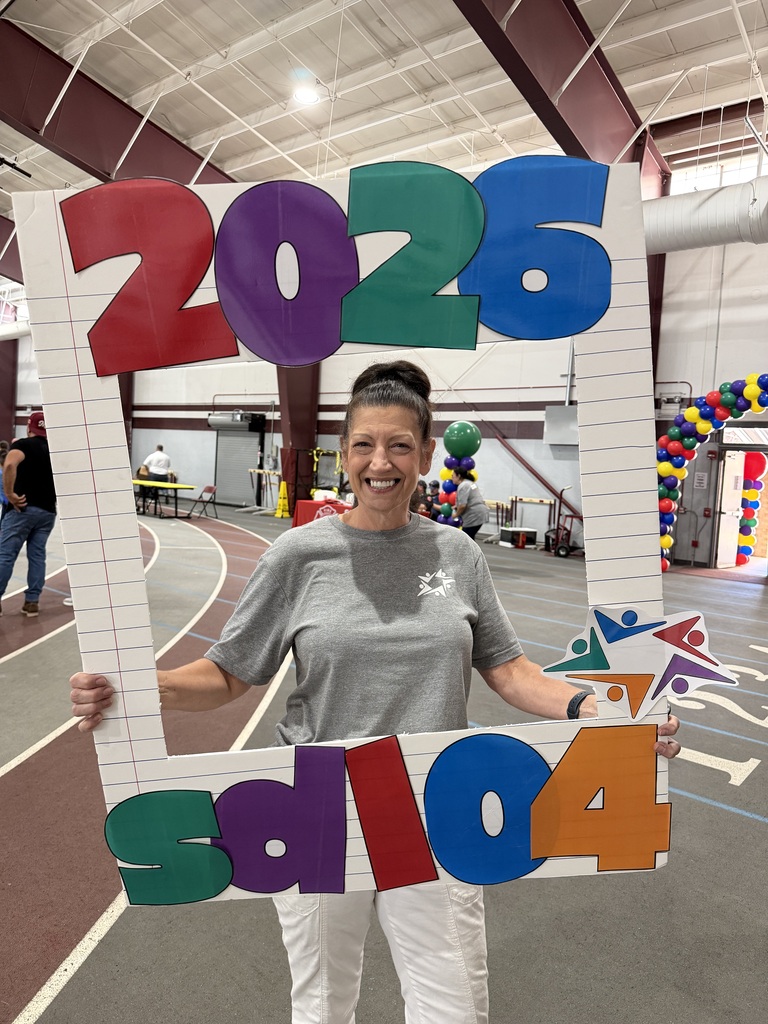 Smiling woman holds a colorful “2025” sign in an indoor gym, surrounded by balloon arches and sports courts; festive, celebratory mood.