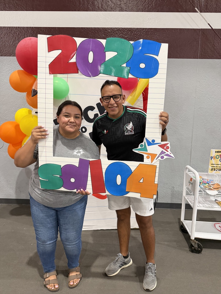 Two smiling people pose behind a colorful “2024” photo frame at an indoor celebration, surrounded by balloons and festive party decorations.