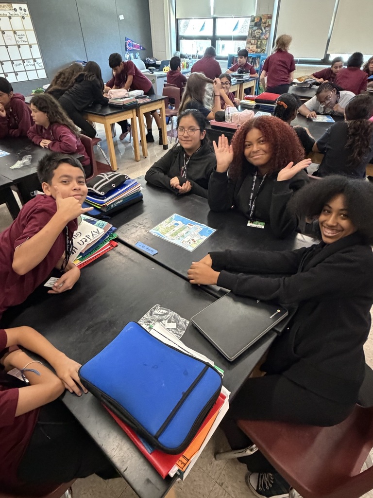 Students collaborate at classroom tables, smiling and waving amid notebooks, binders, and laptops in a lively, welcoming learning environment.