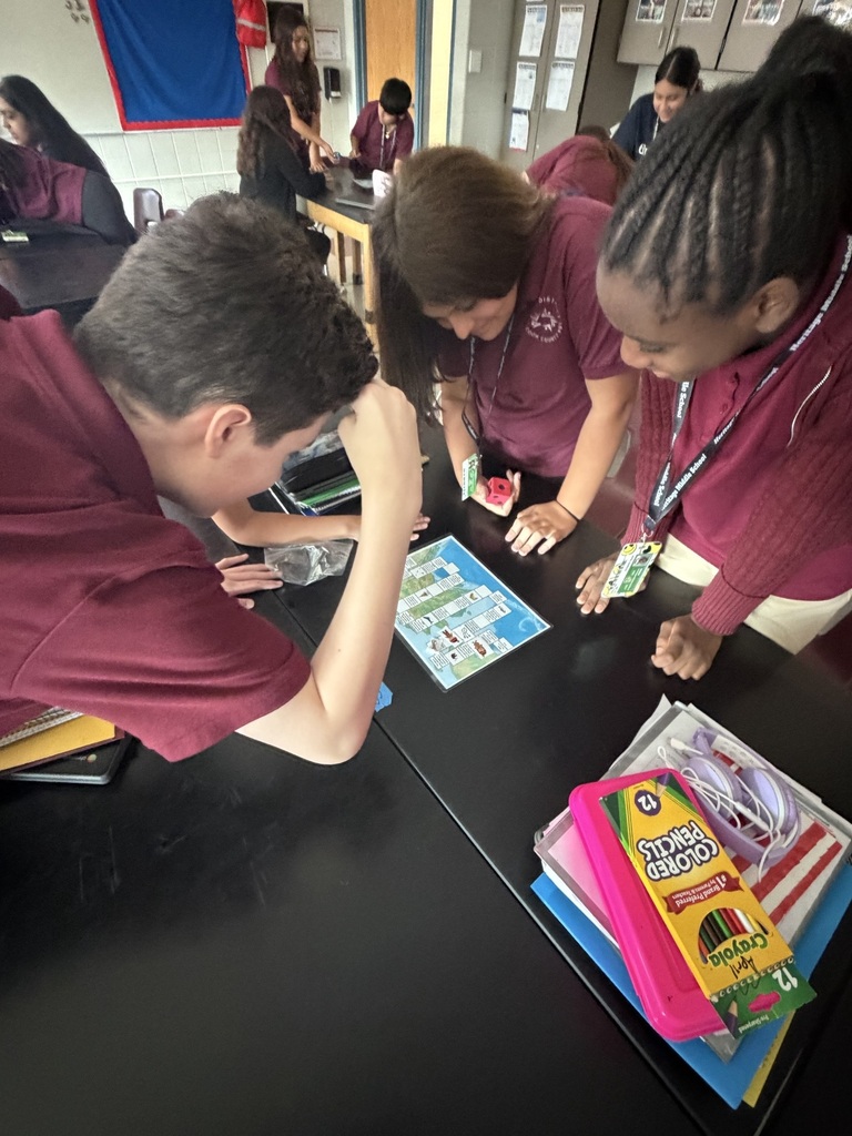 Students in maroon uniforms collaborate around a classroom table, studying a colorful board game and activity materials with focused, upbeat energy.