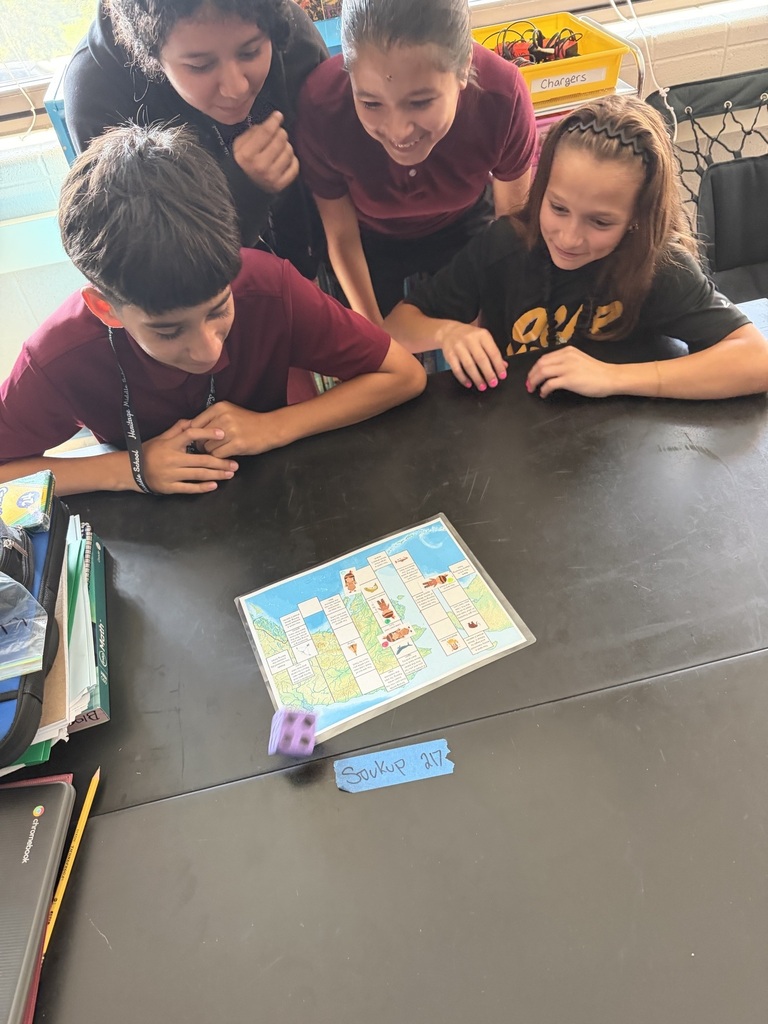 Four children lean over a classroom table, smiling and focused on a colorful board game with dice, sharing an engaged, playful moment.