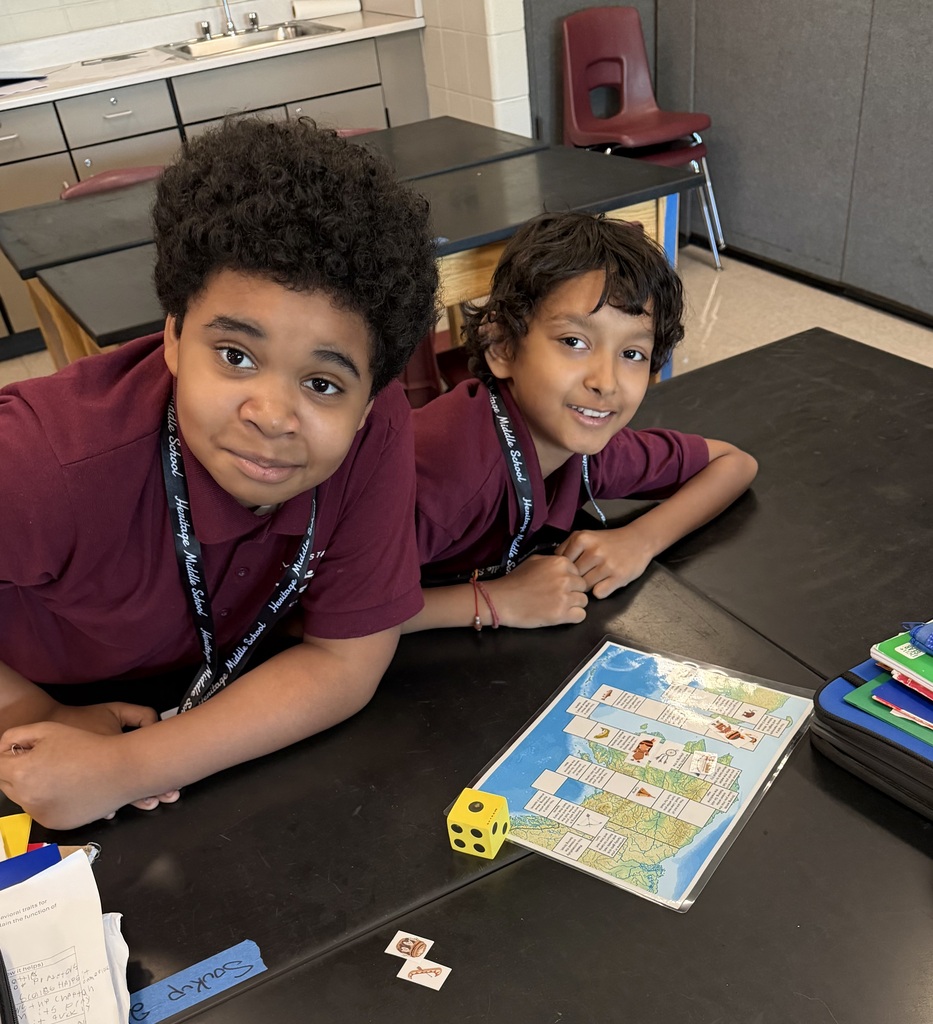 Two smiling students in maroon shirts lean over a classroom table beside a colorful board game, yellow die, and school supplies.