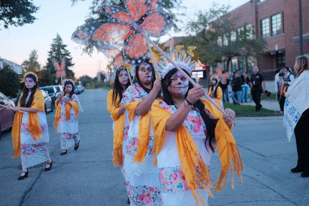 Women in colorful floral dresses and yellow shawls march joyfully in a parade, carrying large butterfly headdresses as onlookers watch.